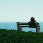 A woman in a hat sits on a bench overlooking the ocean, enjoying a serene summer day.