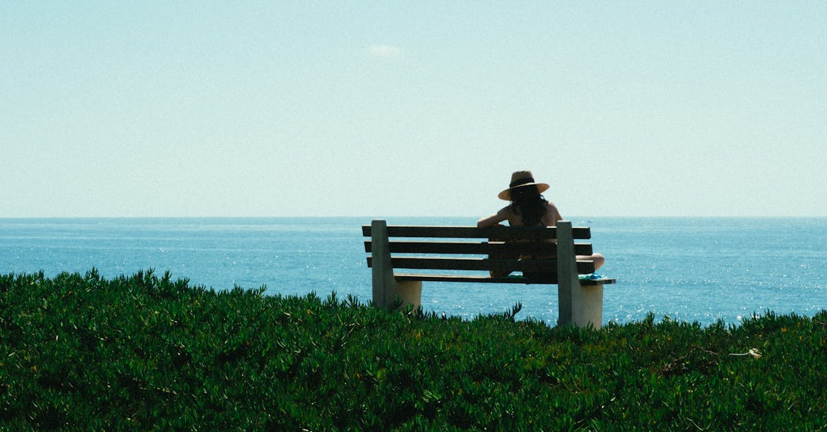 A woman in a hat sits on a bench overlooking the ocean, enjoying a serene summer day.